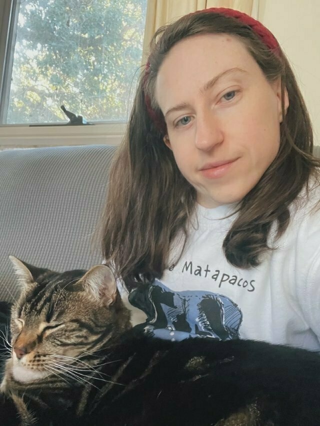 a white girl with long brown hair with a sleepy tabby cat on her lap. the jumper is a bit hard to make out at this angle but you can see illustrated black dog ears and part of its name: ‘Matapacos’