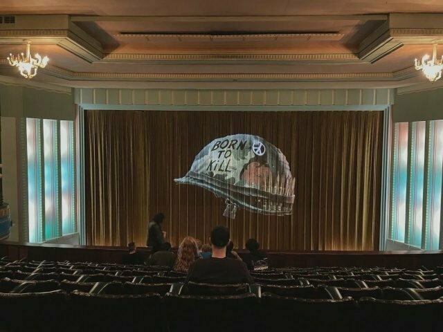 closed curtains in a historic cinema. projected onto the curtains is a military helmet with the words â€œBORN TO KILLâ€� written on it and a peace button attached