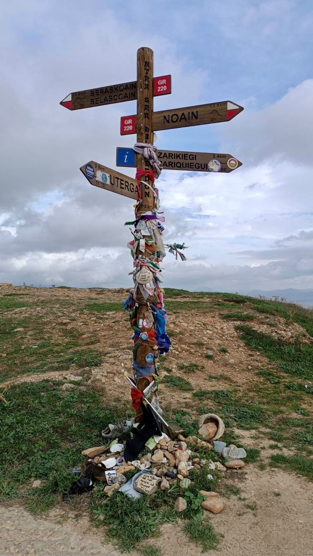 A wooden signpost covered in various objects and ribbons stands at a crossroad against a cloudy sky. Directional signs indicate nearby locations alongside pilgrim mementos.