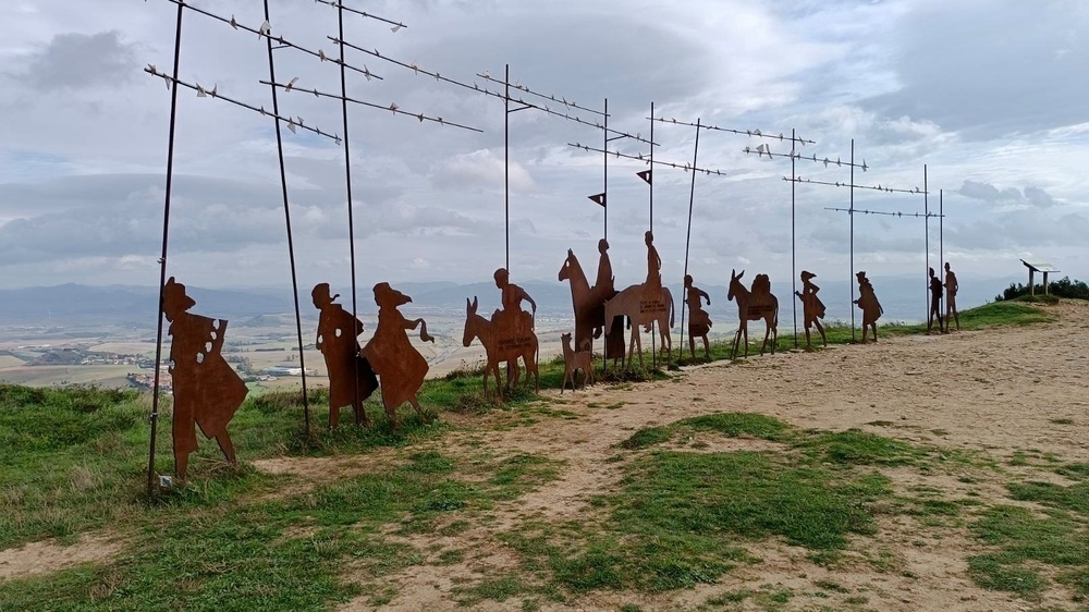 Rusty metal silhouettes of people and animals stand on a hilltop, resembling a procession or historical scene, overlooking a vast, cloudy landscape.