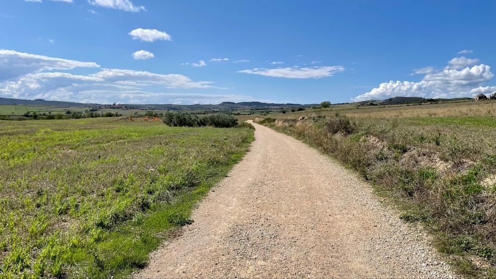 A dirt path stretches through a green landscape with distant hills, under a partly cloudy sky.