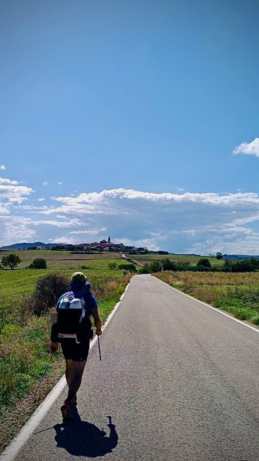 A hiker with a large backpack walks along a road toward a village with visible church spires, set in a rural landscape under a bright, cloudy sky.