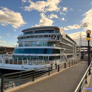A large modern white cruise ship or river boat docked at a waterfront promenade with multiple decks, large windows, and curved architecture, moored alongside a paved walkway with metal railings and ornate lampposts under a dramatic cloudy sky with bridges visible in the background.