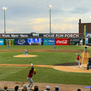 Baseball game in progress viewed through protective netting, showing a pitcher in light blue uniform throwing to a batter in red, with outfielders positioned on the field. Outfield wall displays advertisements for Blaze Credit Union, HOME RUN PORCH, ECOLAB, CHS, TRIA, Coca-Cola, and MORRIE'S with tagline \