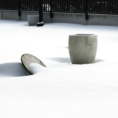 Photograph of a snow-covered patio or terrace with large concrete planters partially buried in deep pristine snow. A barrel-shaped planter stands upright in the centre while a circular lid or shallow dish leans at an angle beside it. A third smaller planter is visible in the background near a black metal railing and concrete wall. The scene is minimalist and nearly monochromatic in white and grey tones.
