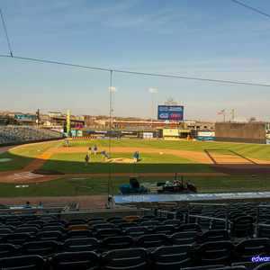 Baseball stadium view from the stands showing an empty field with players in blue uniforms practicing on the infield. CHS FIELD visible on scoreboard with TV & RADIO BROADCASTS sign, CW and KFAN logos. Floodlights illuminate the field during late afternoon with clear blue sky overhead.