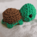 A photo of a small crocheted turtle sitting on an off-white cloth. the turtle has green skin and a brown shell. It is simple and cute with a head almost as big as the rest of the body.