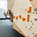 A person wearing climbing shoes is bouldering on an indoor climbing wall with orange and green holds. The climbing facility features padded flooring and additional climbing walls in the background. The climber’s face is covered with a smiley face emoji