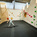 A child standing inside an indoor rock climbing gym, wearing a smiley face emoji over their face. The climbing walls feature various colored holds in blue, yellow, orange, pink, green, and gray. There is a hexagonal wooden structure inset into