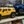 A photo of two toy cars, a yellow SUV and a blue truck, on a wooden shelf with a blurred red and gray background.