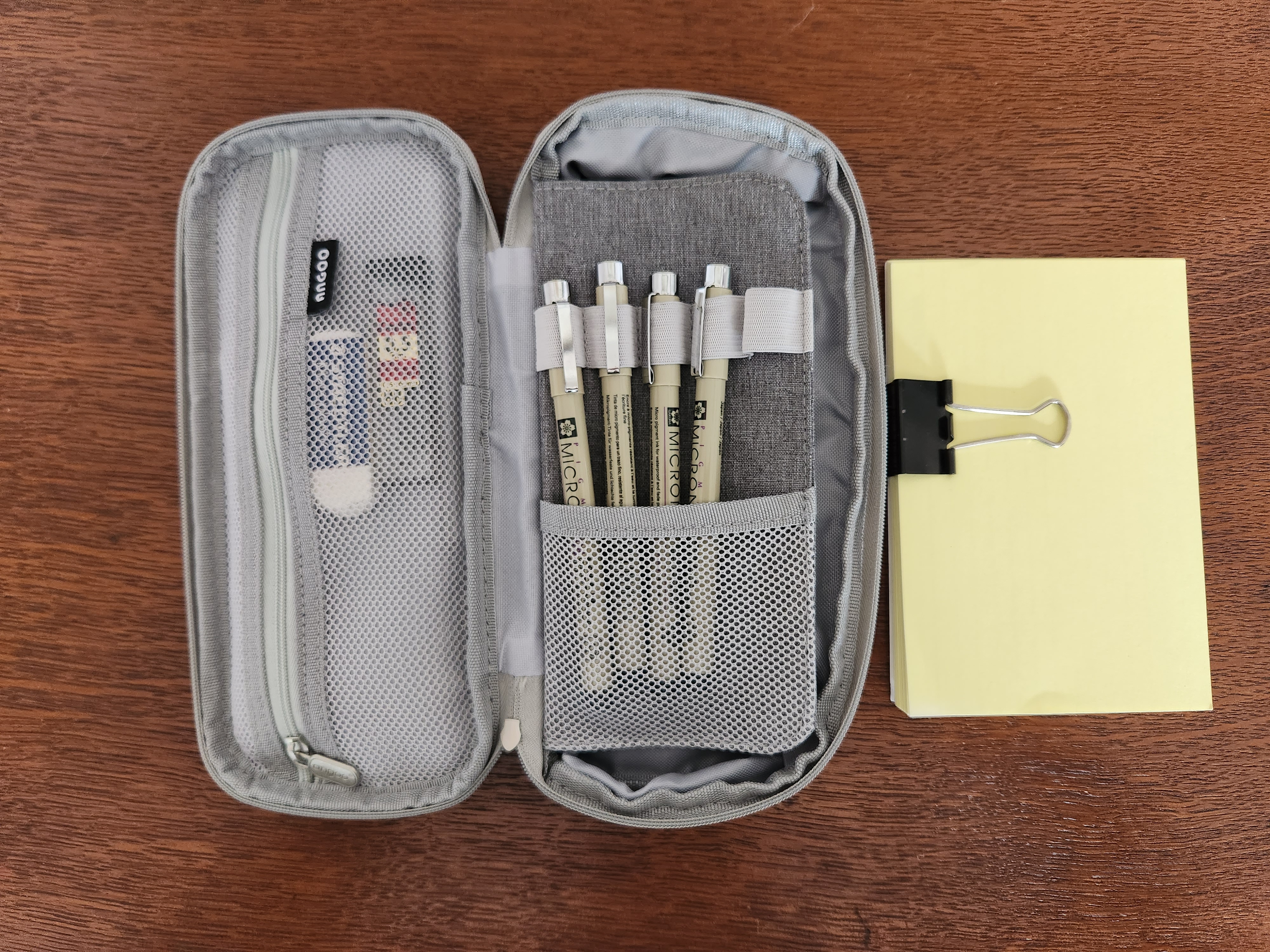 A grey pen case with a zippered pocket on the left, a partition with space for five pens to be held in elastic loops with a mesh net pocket below it. Not pictured, there is a space for more pens or index cards which could be placed under the partition.