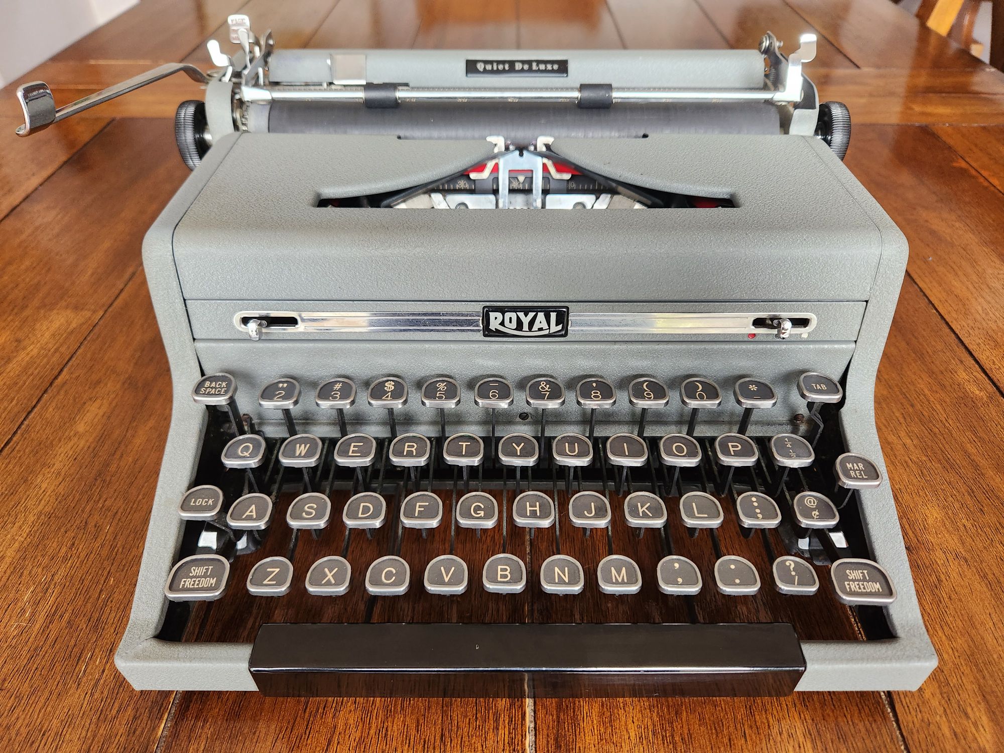 Front view down onto a gray crinkle painted Royal Quiet De Luxe with black glass keys and a wide black space bar which overlays the metal frame of the front of the machine. The typewriter sits on a polished wooden table which provides a stark natural contrast to the industrial nature of the typewriter's esthetic.