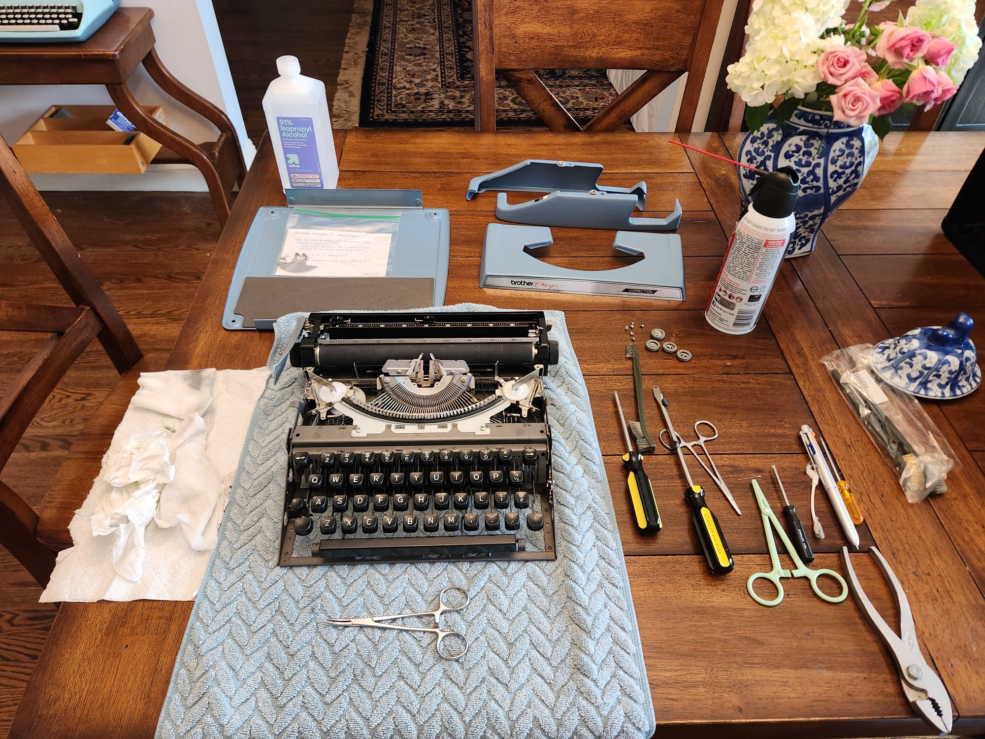 Wooden table with a blue towel on top of a portion. On top of that is the internal frame and components of a typewriter with the hood, bottom, and side piece of the machine sitting behind it. Strewn around it are a variety of screwdrivers and small tools as well as a can of compressed air.