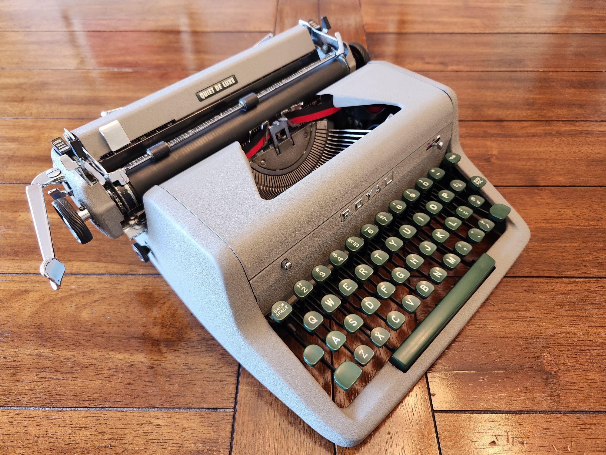 View down onto a 1955 Royal Quiet De Luxe Portable Typewriter with a gray body and green keys sitting on a polished wooden table.