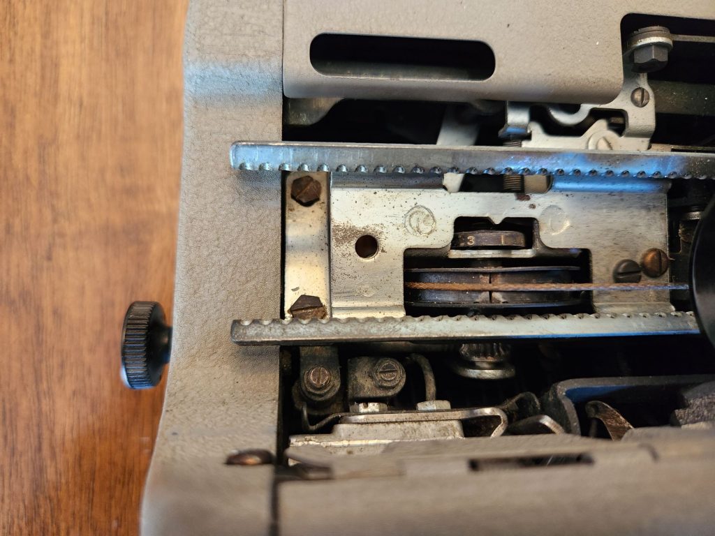 View down into the left side of the typewriter carriage on a 1955 Royal Quiet De Luxe. On the outside of the machine on the far left is a black thumbscrew knob which attaches to an unseen metal rod which actuates a gear that attaches through the hub for the typewriter's drawband just above the gear. Just above this is a small indicator wheel which appears to be set at about 3.5.
