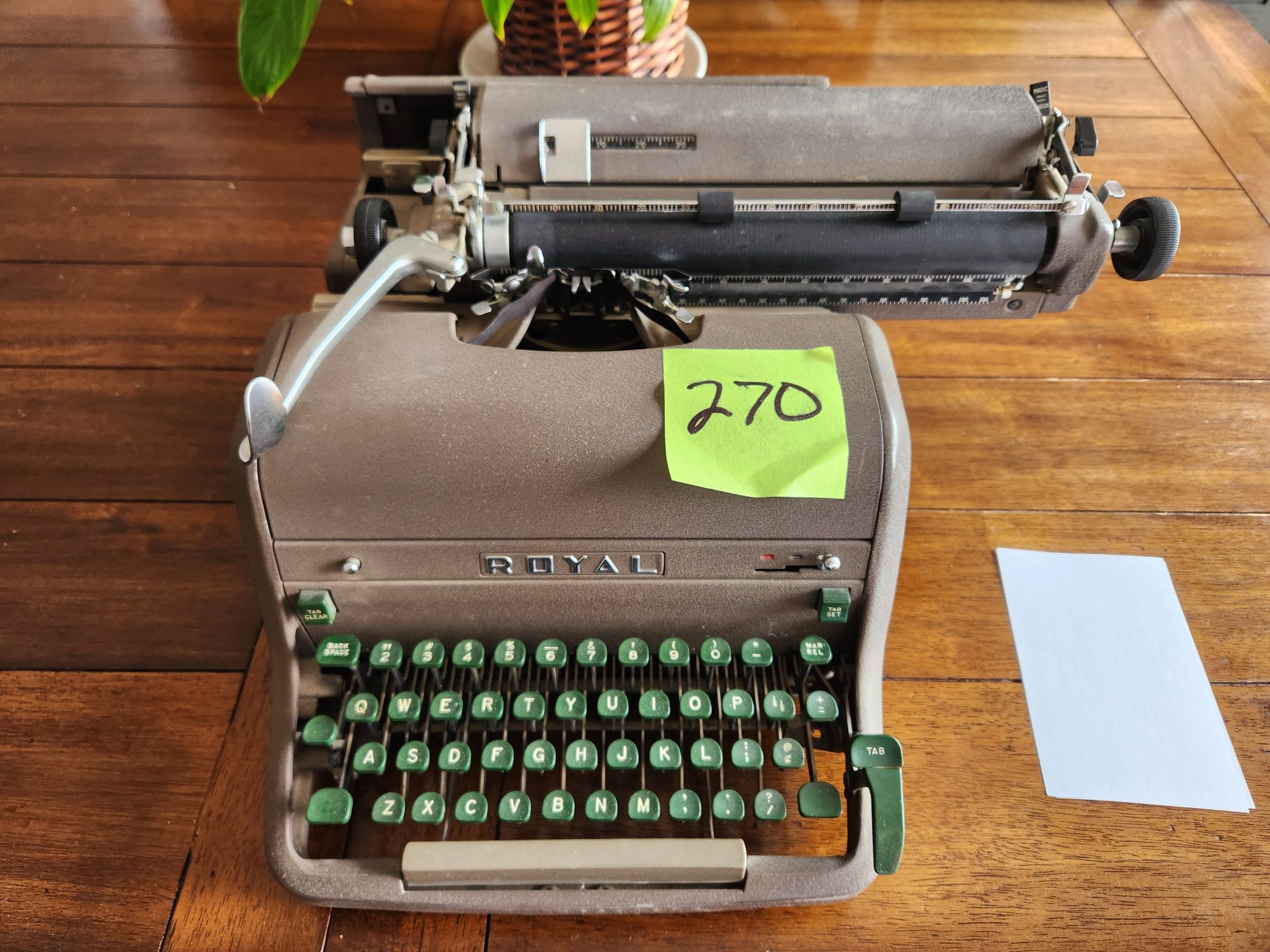 A Brownish gray 1955 Royal HH typewriter with green plastic keys sits on a wooden plank table. There's a yellow post it note on the right side of the hood with the Sharpied number 270 on it. Of note, the right side of the keyboard has a long tab button to be actuated by the side of ones right hand rather than tapped by a finger.