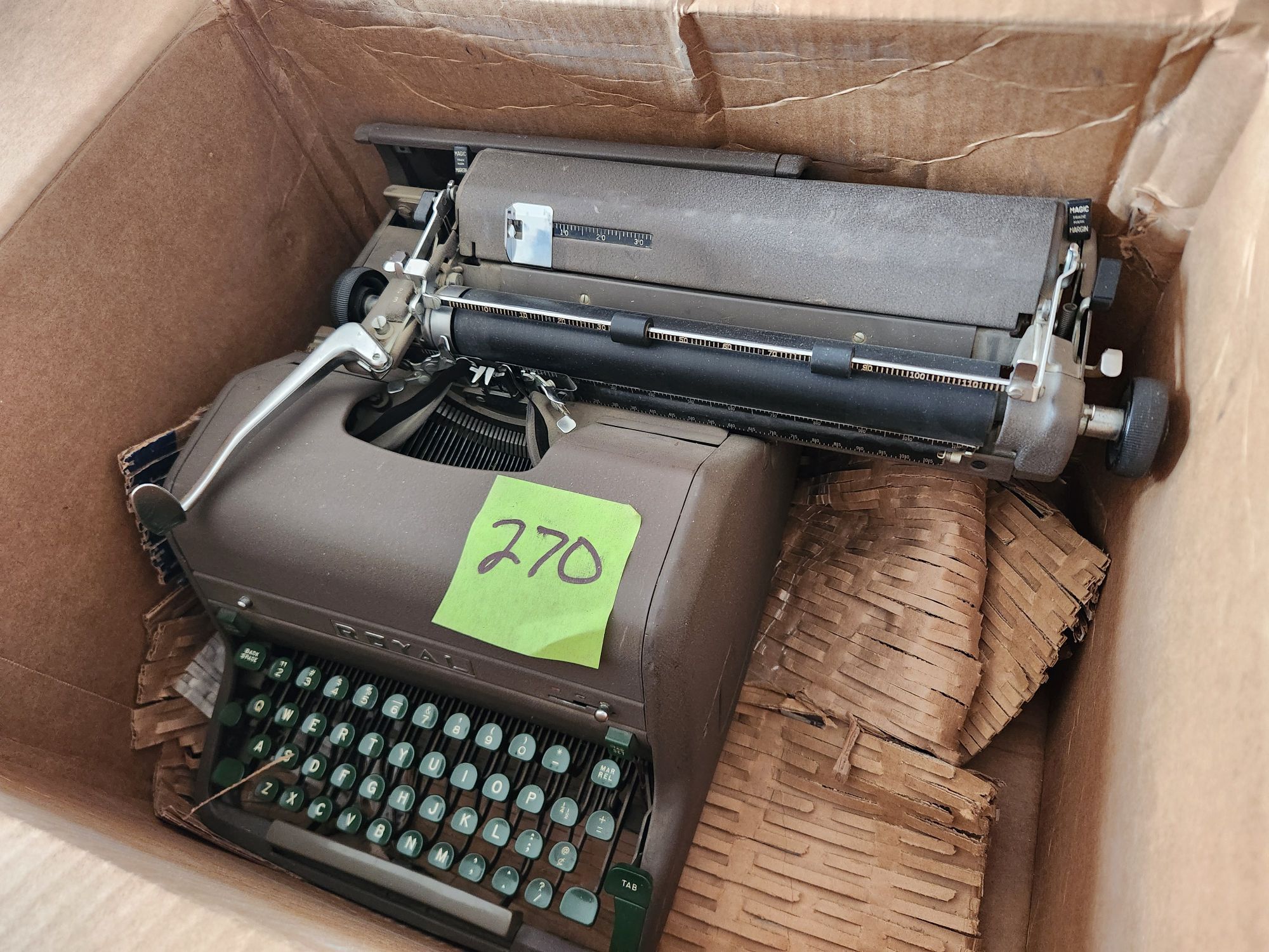 A Royal HH typewriter sitting in the left side of a box that's too short front to back and too wide left to right. The carriage on the machine is all the way to the right. 