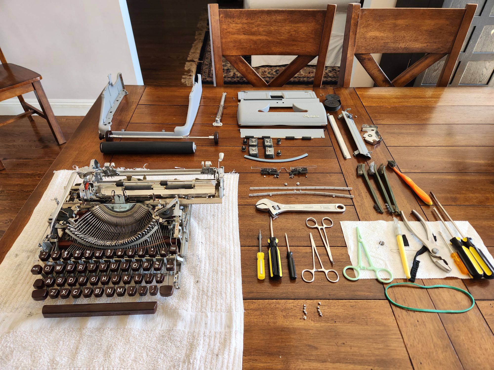 A busy photo of a deconstructed Olympia SM3 typewriter sitting in pieces on a wooden dining room table. The skeleton of the machine and keys are on a white towel in the front with the platen, rod, and knobs behind it. Also in the background are the shell of the body, the paper table, and measurement guides. To the side are a variety of screwdrivers, wrenches, brushes, and miscellaneous tools. Small piles of screws are near the pieces from which they were removed.