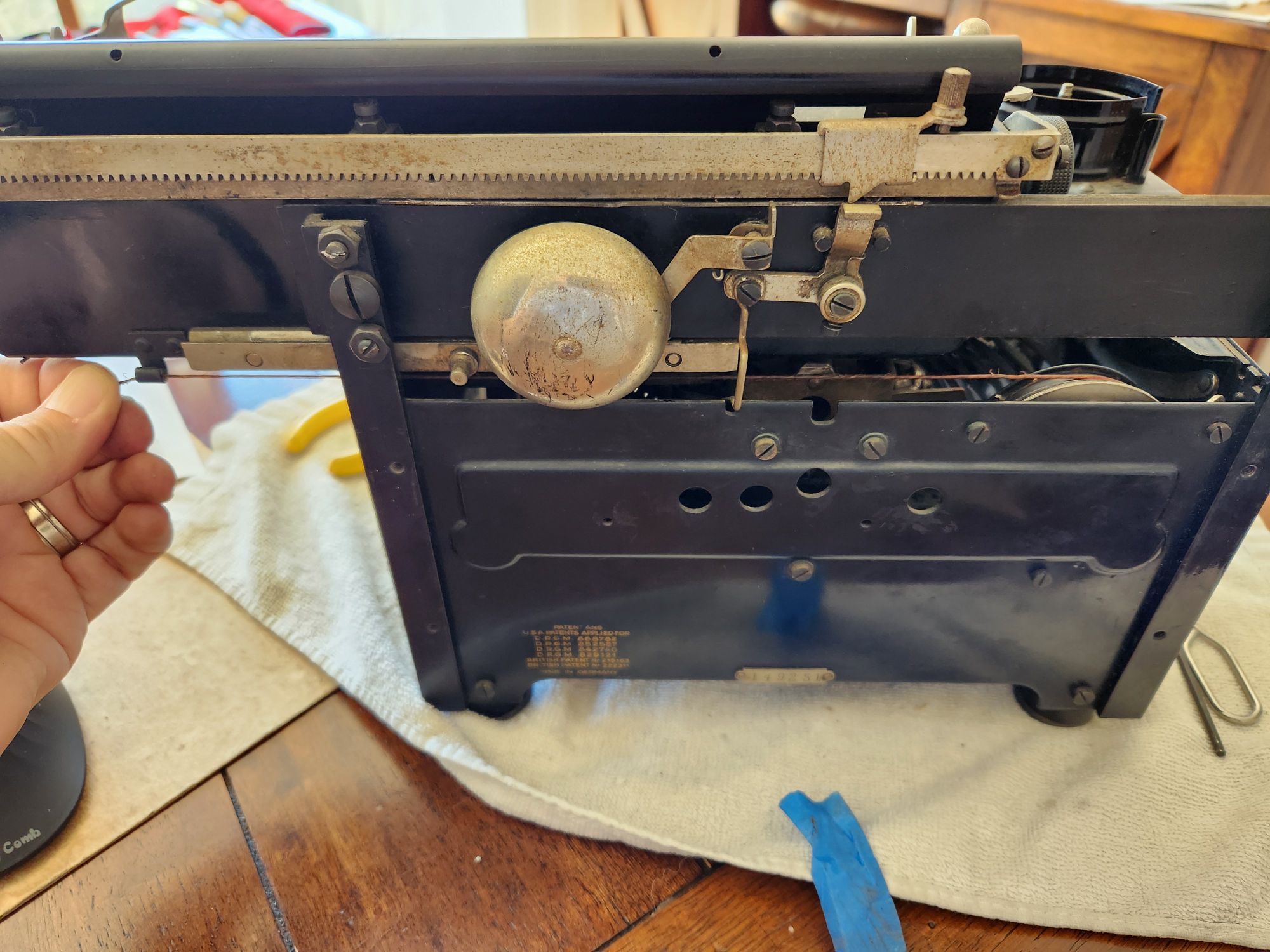 Rear of an Orga Privat 5 typewriter featuring a hand at the left side holding the drawstring where it needs to be attached to the carriage. On the other side the string is attached to the mainspring wheel which has a thin silver knurled ratchet system attached to it.