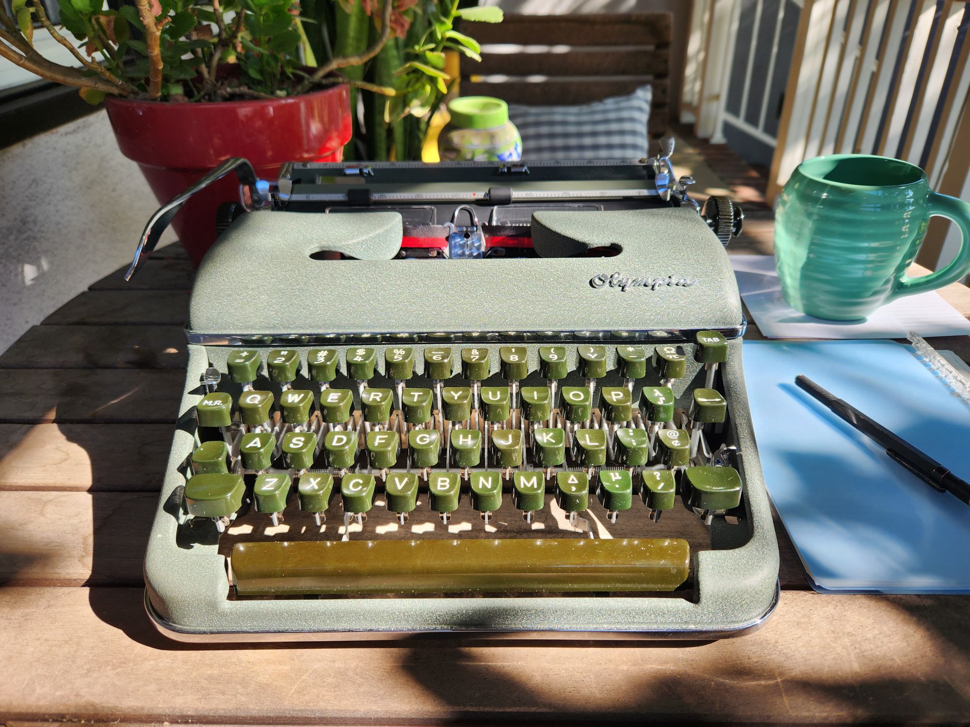 A green crinkle painted Olympia SM3 with chrome highlights, green plastic keys, and a greenish-brown space bar sits on a sun dappled table next to a small potted plant. Off to the side are a small notebook, mechanical pencil and green coffee mug creating a very cozy morning atmosphere.