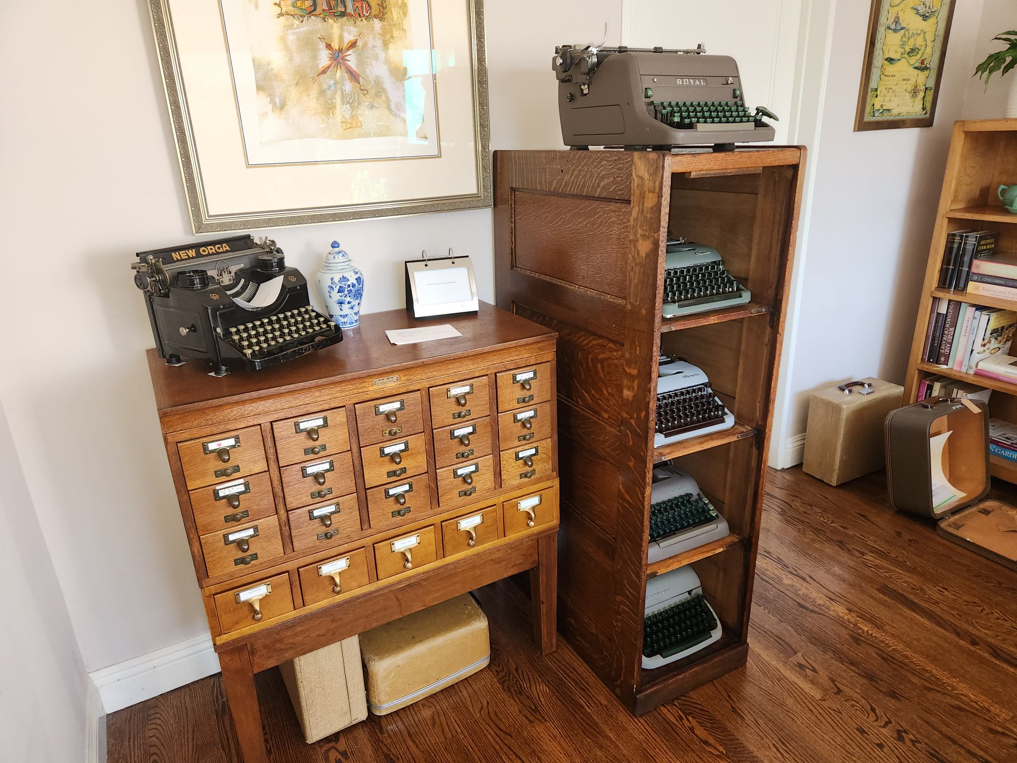 A Fred Macey Company 20-drawer card index filing cabinet with the drawers and hardware removed. In their place we see four typewriters peeking out from each of four shelves inside and an additional typewriter on top.  Next to the filing cabinet is a Gaylord library card catalog with a black typewriter on top along with some additional decorations. 