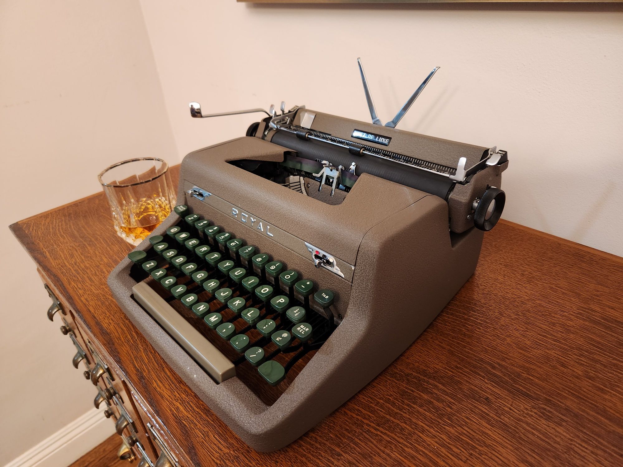 View of a 1952 Royal Quiet De Luxe in brown frieze paint and green plastic keys sitting on a wooden library card catalogue. An old fashioned glass of bourbon sits in the background.