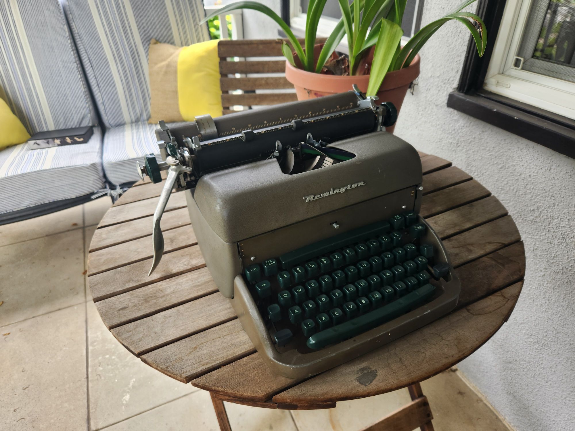 Angle down on a 1951 Remington Super-Riter standard typewriter in a military green crinkle paint with green plastic keys. It sits on a round wooden bistro table in a porch setting.