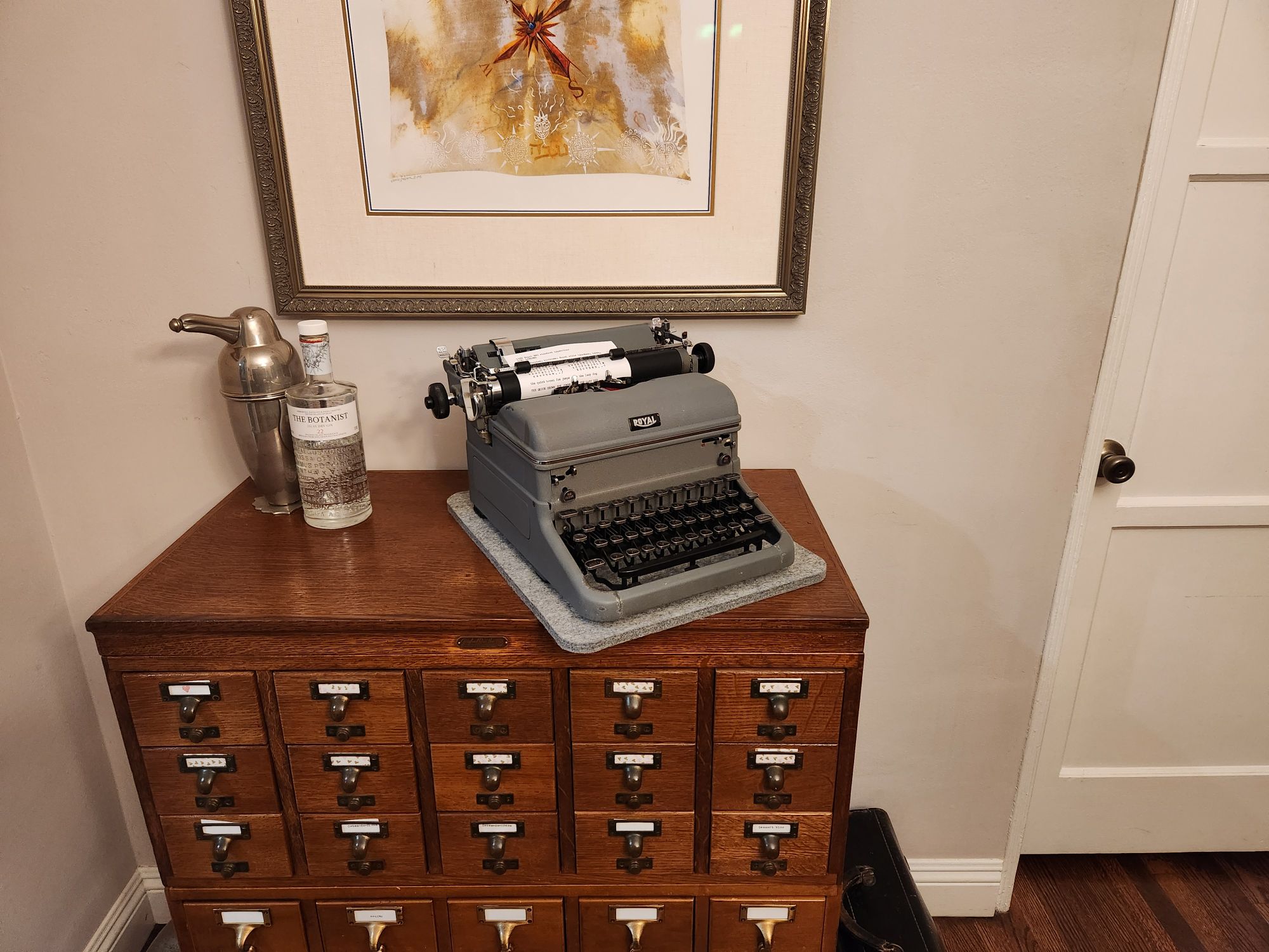 Hero view of a 1950 Royal KMG standard typewriter sitting on a library card catalog next to a bottle of gin and a penguin-shaped cocktail shaker