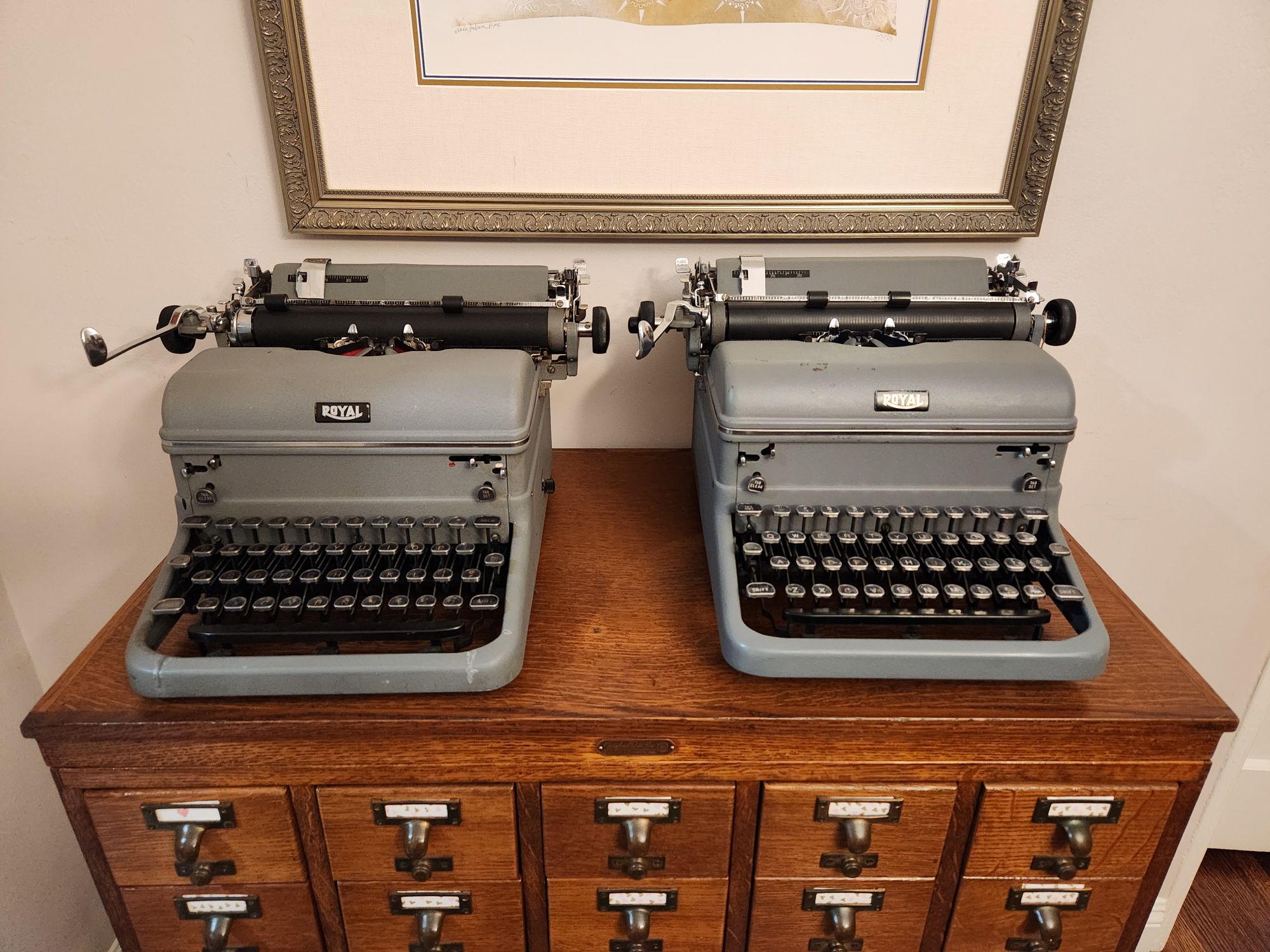 Twin gray frieze 1950 Royal KMG standard typewriters sitting next to each other on a wooden library card index. Careful inspection will show one to be in elite and the other in pica.