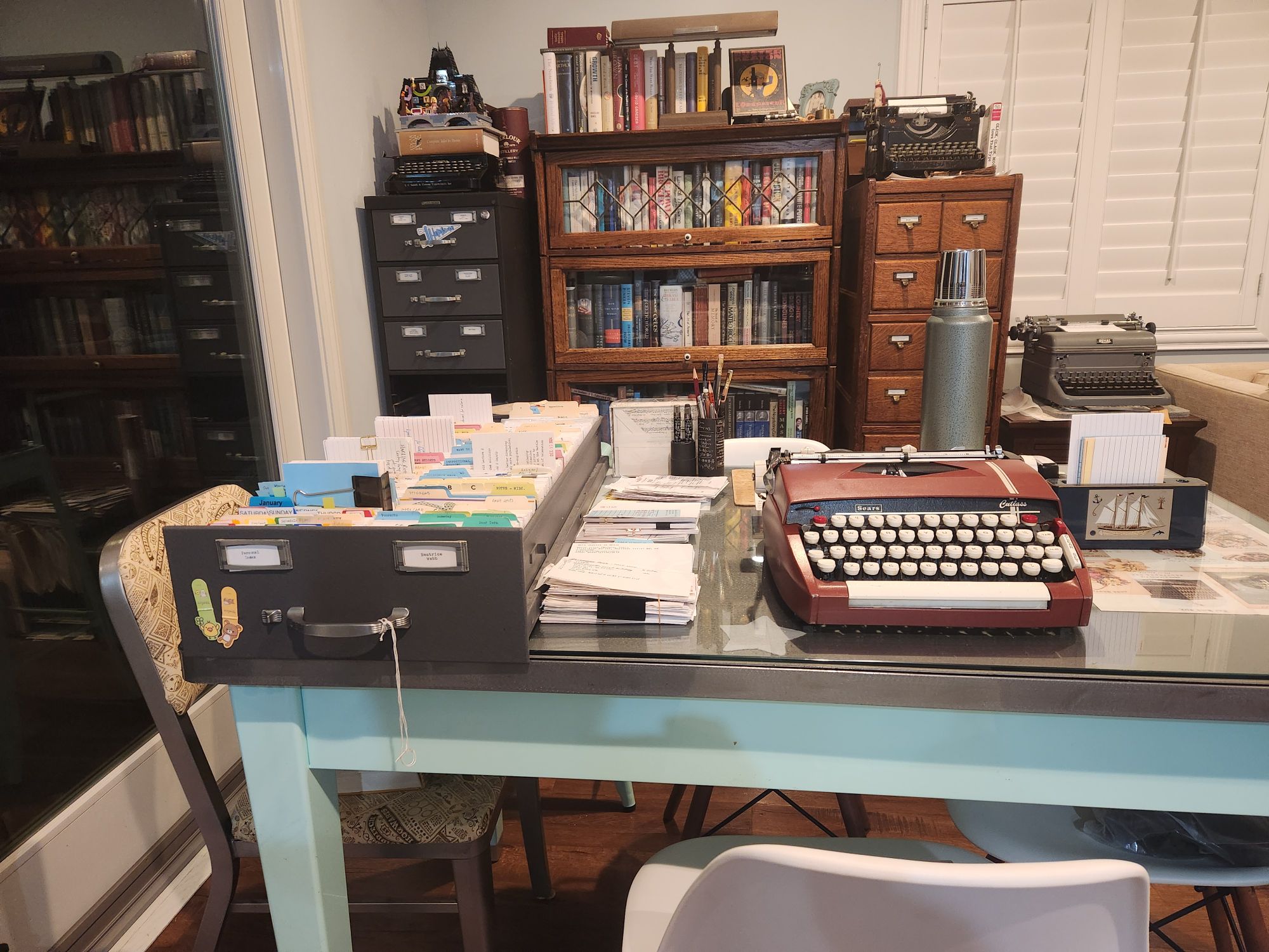 A steel desk with several card index filing cabinets and a bookcase in the background. On the desk next to a typewriter is a double drawer of 4x6