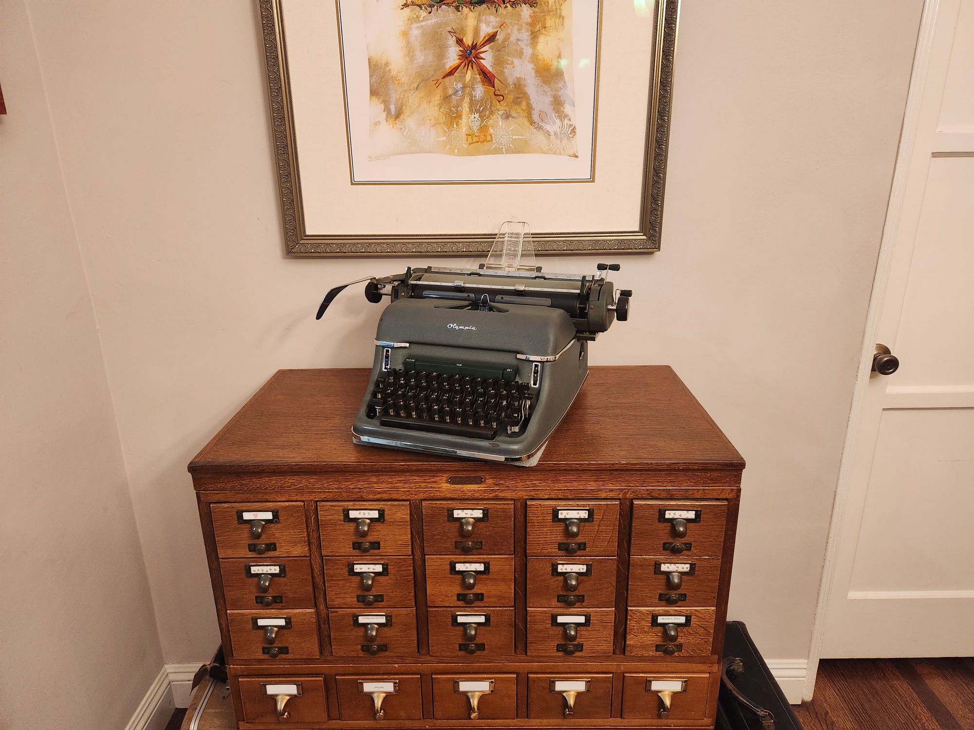 A green crinkle painted 1956 Olympia SG1 standard typewriter sits at an angle on top of an oak library card catalog filing cabinet