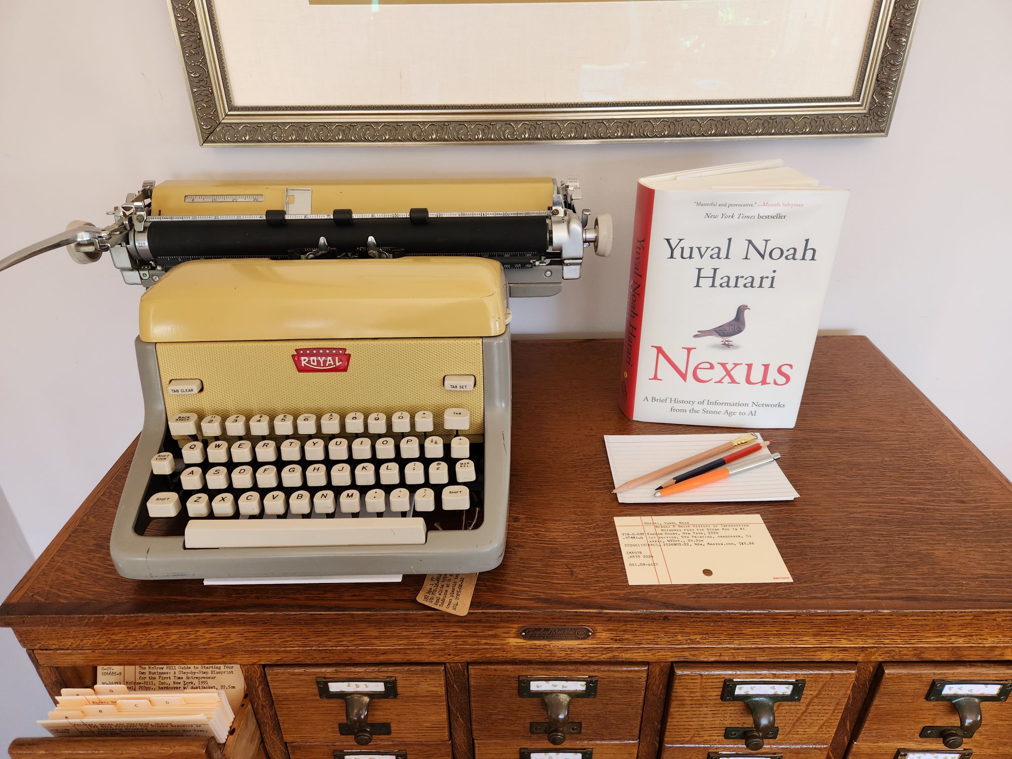 A copy of Harari's book Nexus standing next to a sandstone yellow Royal FP typewriter with index cards sitting in front of it with several pencils and a pen. They all sit on top of an oak library card catalog.