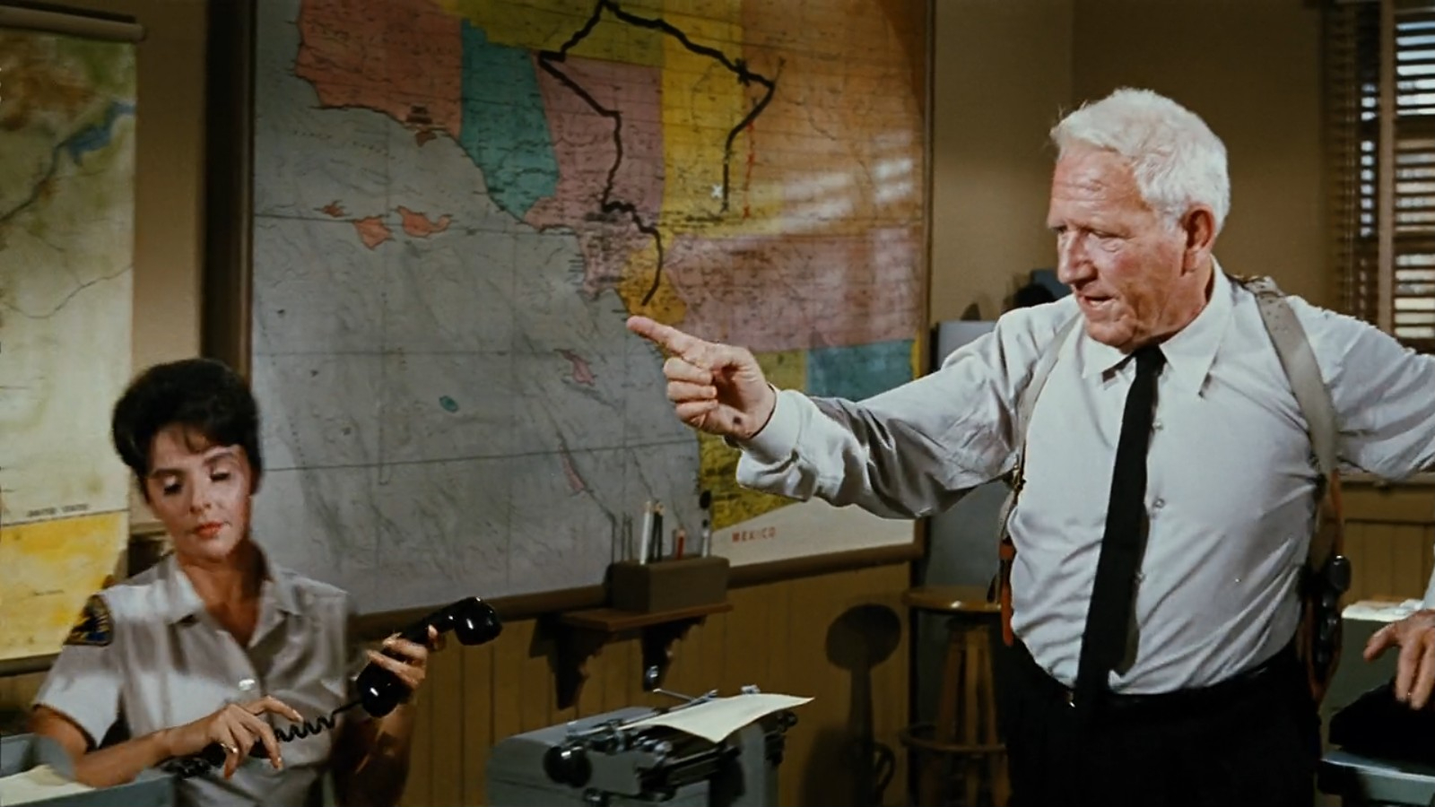 Tracy pointing across the room in the foreground as his secretary types on a standard Royal KMG.