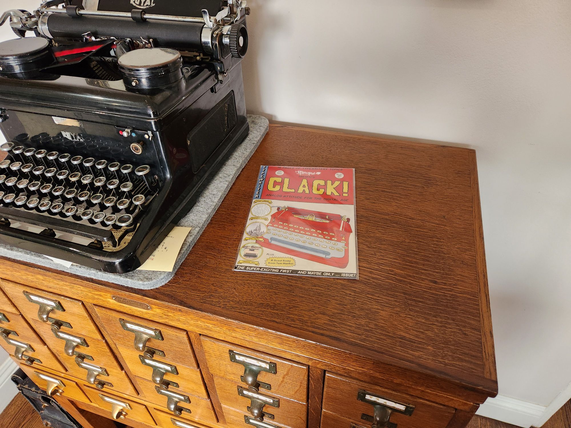 Issue No. 1 of Clack!, a colorful zine in red, yellow, and blue with a cartoon-like illustration of a Corona flattop typewriter on the cover sits on top of a 20 drawer library card catalog next to a black lacquer Royal KHM standard typewriter