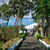 Tree-lined path leading to a yellow wooden building and gated entrance, with tropical palms and a coastal view beyond