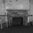 A brick fireplace with a weathered wooden mantelpiece in a room with peeling paint and two old wooden chairs