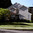 Two-storey suburban house with brick and grey vinyl siding, arched window, manicured lawn, and mature trees on a corner plot
