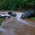 Muddy river cascading over rocks into a swirling stream, surrounded by dense green woodland