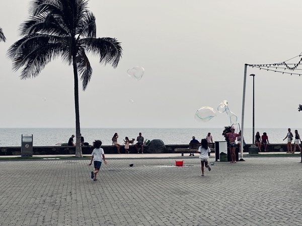 Kinder spielen mit Seifenblasen an der Strandpromenade von Maspalomas, während Leute auf einer Mauer sitzen, und Palmen stehen im Hintergrund.