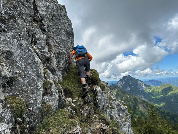 Eine Person mit einem blauen Rucksack klettert einen felsigen Bergpfad hinauf, umgeben von einer malerischen Berglandschaft.
