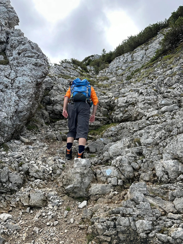 Eine Person mit einem blauen Rucksack klettert einen steinigen Bergpfad hinauf.