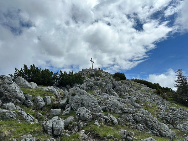 Ein Gipfelkreuz steht auf einem felsigen Hügel unter einem teilweise bewölkten Himmel.