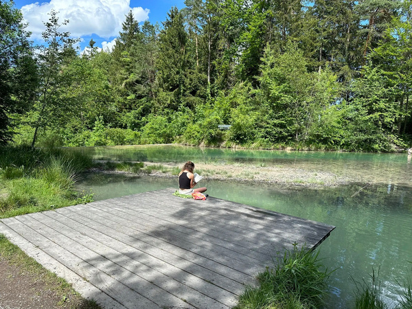 Eine Person sitzt auf einem Holzsteg an einem ruhigen, von Bäumen umgebenen See.