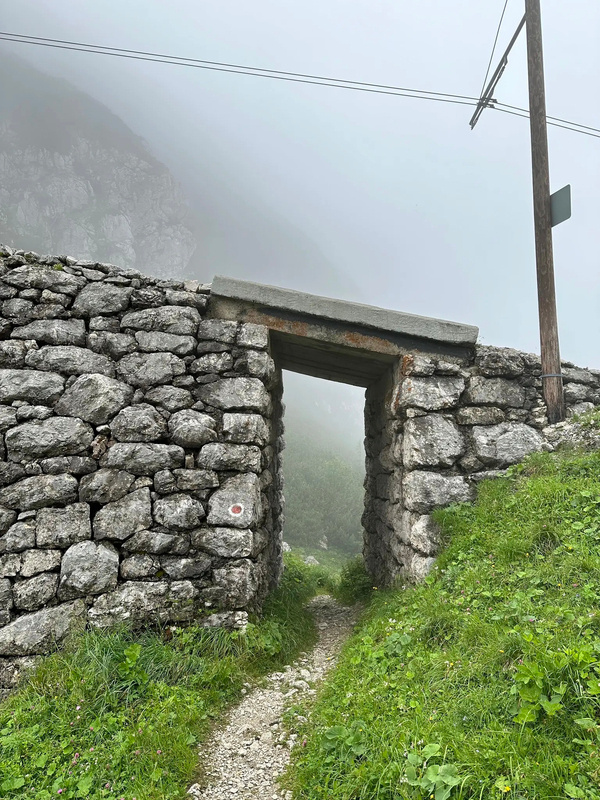 Ein schmaler Pfad führt durch eine Steinmauer in einer nebligen, bergigen Landschaft.