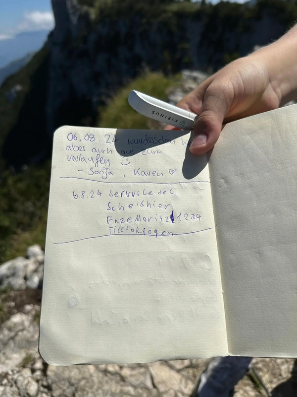 Eine Hand hält ein aufgeschlagenes Notizbuch mit handgeschriebenen Einträgen, während im Hintergrund bergige Landschaft zu sehen ist.
