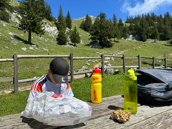 Auf einem Holztisch sitzt ein Kind mit Baseballkappe neben einer Brotzeit und Getränken in einem malerischen alpinen Landschaftshintergrund.