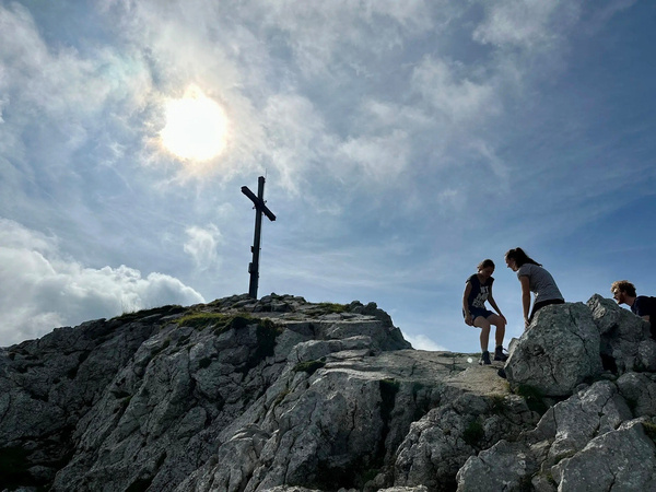 Drei Menschen sitzen auf einem felsigen Berggipfel neben einem großen Gipfelkreuz, während die Sonne hinter Wolken hervorschaut.