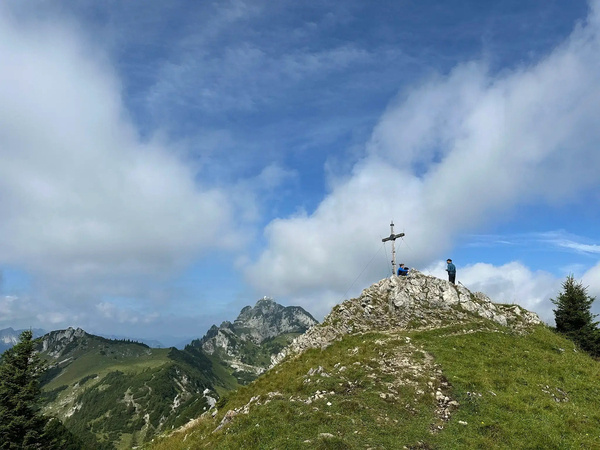 Ein Wanderer steht neben einem Gipfelkreuz auf einem felsigen Hügel mit einer weiten Berglandschaft im Hintergrund unter einem bewölkten, blauen Himmel.