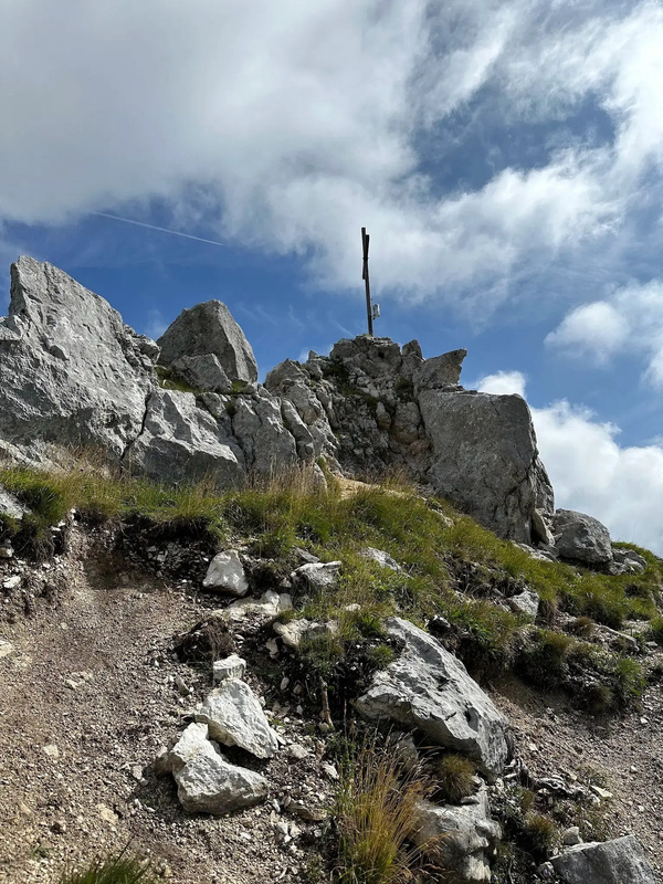 Ein Gipfelkreuz steht auf einem felsigen Berggipfel, umgeben von Felsen, Gras und wolkigem Himmel.