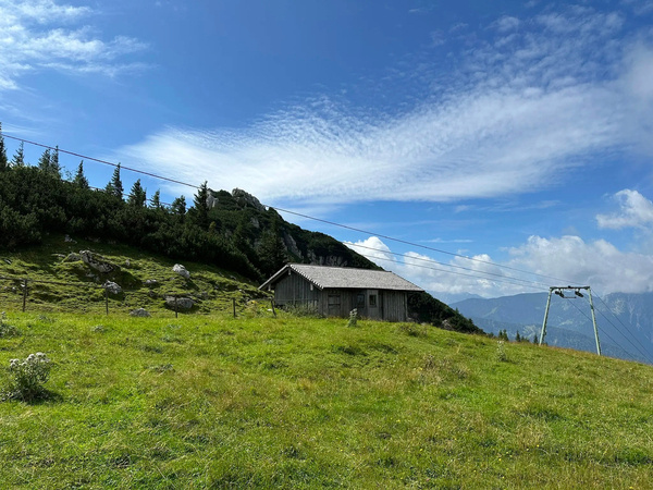 Eine Berghütte steht auf einer grünen Wiese vor einem bewaldeten Hang und einem klaren blauen Himmel.